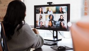 Back of a businesswoman in her home office meets with colleagues on a web conference, with a grid of faces on her computer.