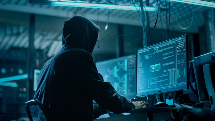 A person wearing a hood uses a computer in a dark room.