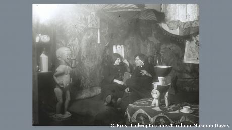 The photo shows Ernst Ludwig Kirchner sitting in his studio with his partner Erna Schilling. The two are surrounded by sculptures, furniture and heavily patterned tapestries