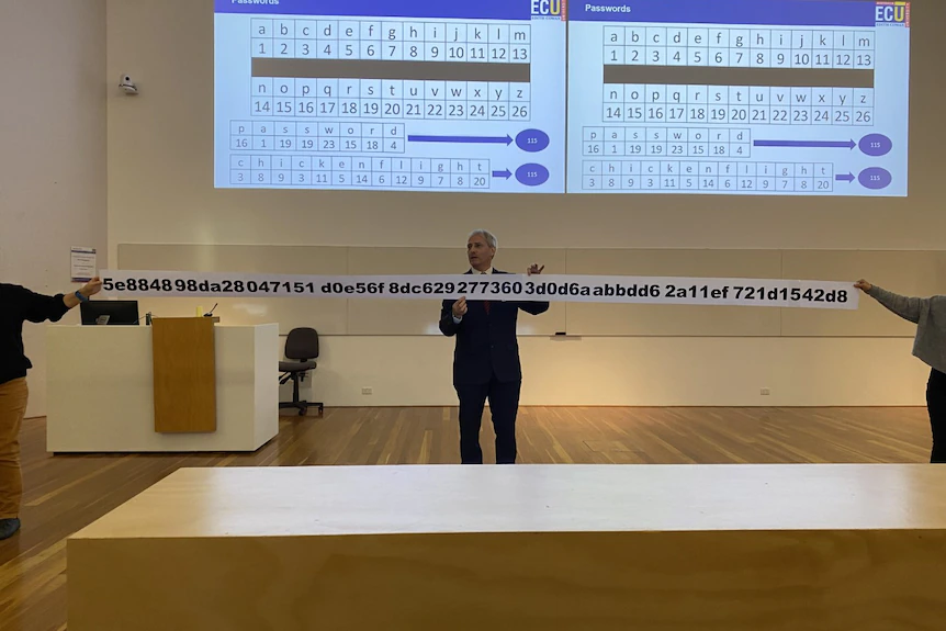 A man in a suit giving a lecture holds a ridiculously long piece of paper containing a password of random letters and numbers.