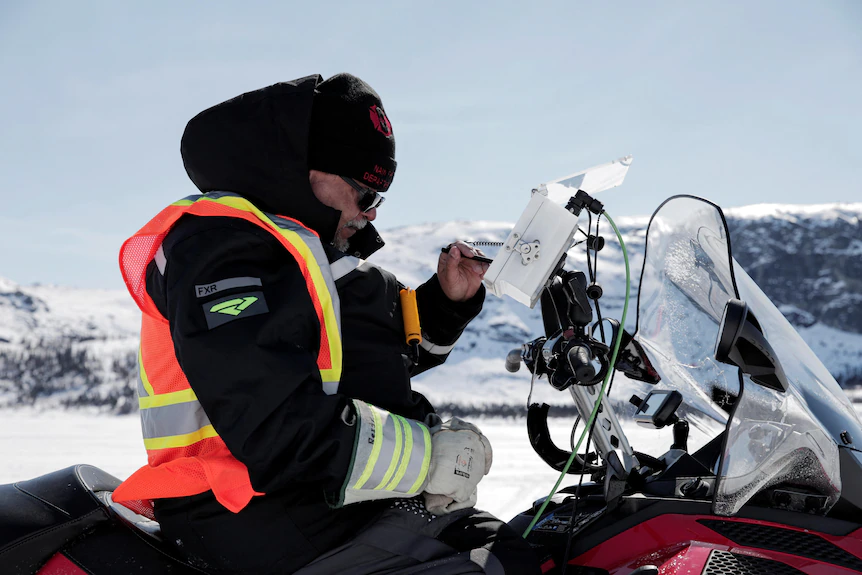 A man wearing thick clothes and high vis, is sitting on his snowmobile, looking at a small screen to check data.