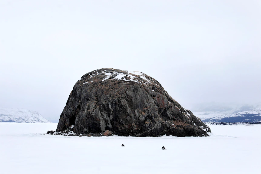 A very large dark rock is pictured in a white, snowy environment. In front of it two very small people driving snowmobiles.
