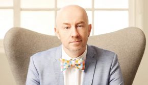 A bald man in a bow tie sits on a couch.