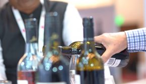 A close up of a wine being poured, other wines with screw tops,  man with a lanyard in blurred foreground.
