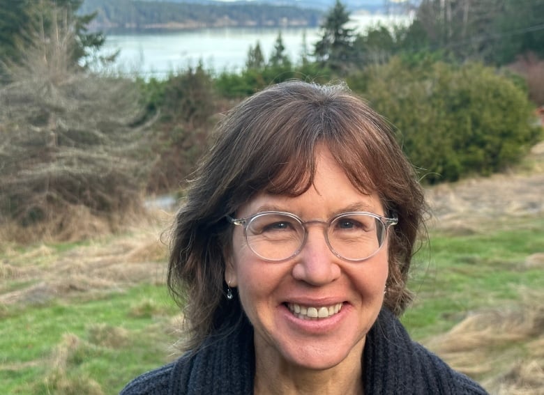 A smiling woman in glasses and a black sweater stands outdoors, with a forest, waterway and mountains in the distance behind her.