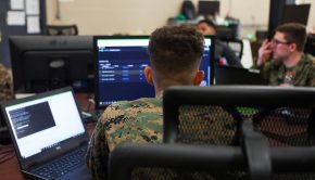A group of Marines work at desks with computer monitors.